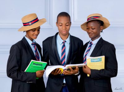Three students in black blazers and straw hats reading open books together.