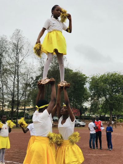 Two students lifting a cheerleader in a yellow skirt above their heads on the sports field.