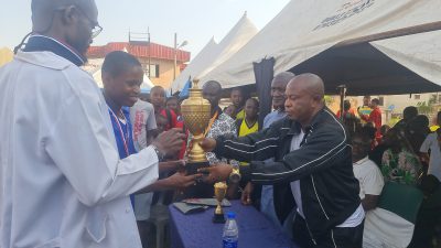 A student in a white shirt and blue lanyard presenting a gold trophy to a man in a black jacket.