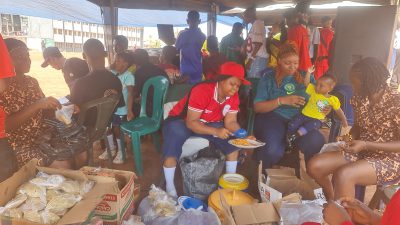 Students seated on green plastic chairs eating from plates during a break in the games.