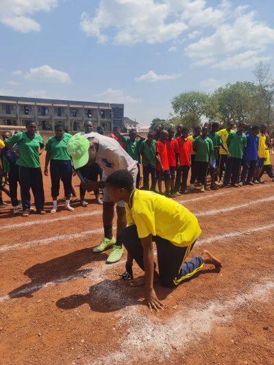 A runner in a yellow vest crouches at the starting line with the green house team lined up behind.